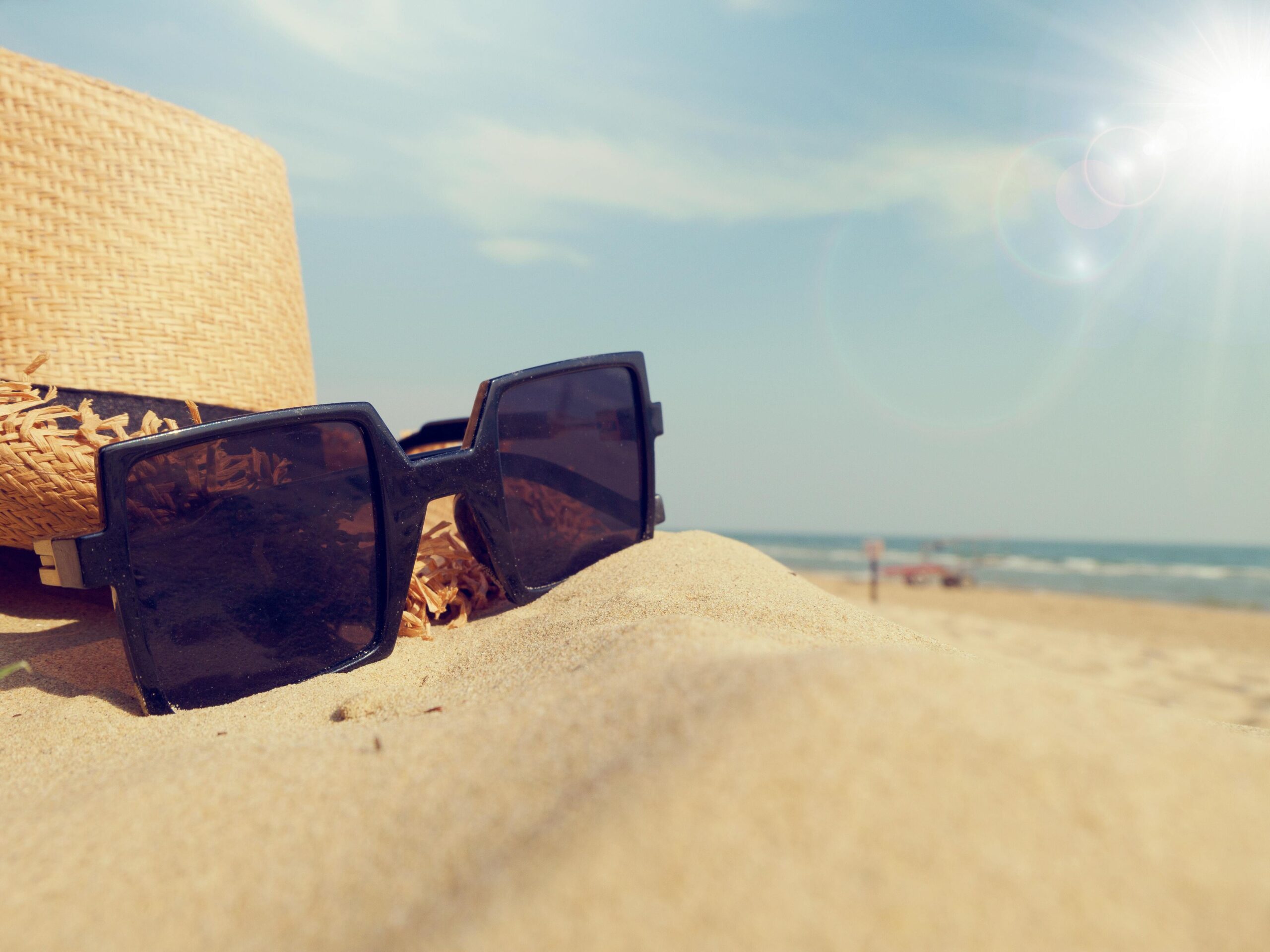 Close-up of sunglasses and straw hat on sandy beach under bright sunlight, perfect summer vibe.